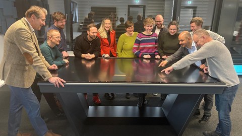 Mehrere Menschen stehen um einen Tisch herum, sehen gut gelaunt aus und zeigen auf eine Grafik auf dem Tisch.