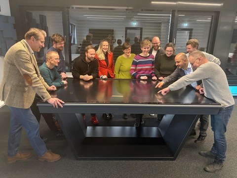 Mehrere Menschen stehen um einen Tisch herum, sehen gut gelaunt aus und zeigen auf eine Grafik auf dem Tisch.
