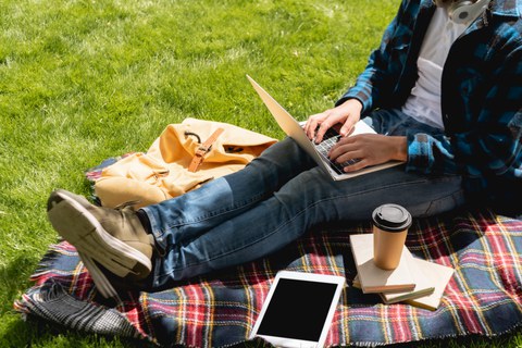 Das Foto zeigt eine studierende Person mit Laptop im Anschnitt. In der Nähe liegen ein Tablet mit leerem Bildschirm und Pappbecher auf einer karierten Decke.