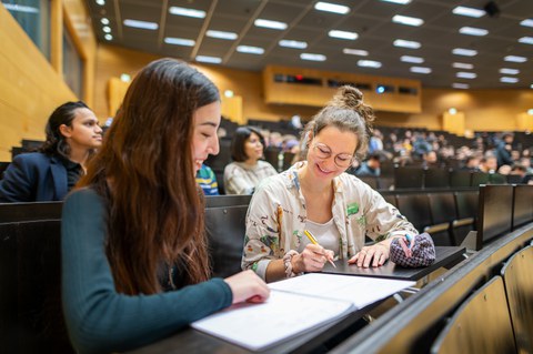 Foto von zwei Studierenden an der TUD, die in einem vollen Hörsaal zusammen arbeiten.