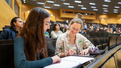 Foto von zwei Studierenden an der TUD, die in einem vollen Hörsaal zusammen arbeiten.