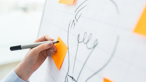 Das Foto zeigt ein Flipchart. Auf dieses wurde eine Glühbirne gemalt. Um die Glühbirne herum kleben leere gelbe Notizzettel. Eine Person schreibt gerade mit einem schwarzen Stift etwas auf einen dieser Notizzettel.