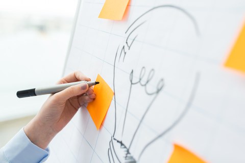 Das Foto zeigt ein Flipchart. Auf dieses wurde eine Glühbirne gemalt. Um die Glühbirne herum kleben leere gelbe Notizzettel. Eine Person schreibt gerade mit einem schwarzen Stift etwas auf einen dieser Notizzettel.
