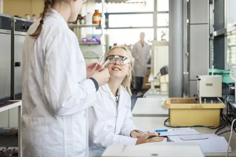 Zwei Frauen unterhalten sich im Labor. Eine davon hält zwei Reagenzgläser in der Hand.