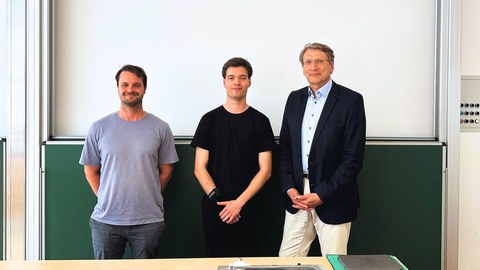 Luis Wunder, Richard Stephan und Prof. M. Beckmann, nebeneinander stehend vor einer Tafel nach der Verteidigung der Arbeit.