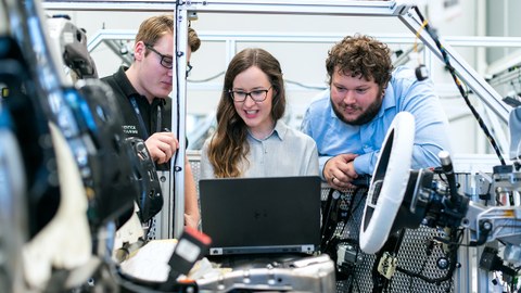 Drei Personen inmitten technischer Apparaturen und Versuchsanlagen, die auf einen Laptop in der Mitte des Bildes schauen.
