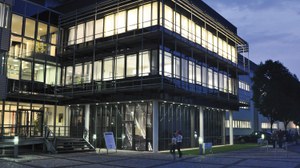 Ths is a photograph of the entrance section of the Fraunhofer IKTS. The night draws in, but all windows are brightly lit, and people are walking in front of the building.