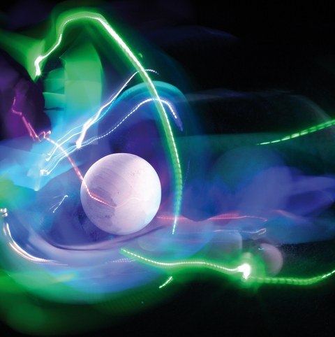 The picture shows an artistic arrangement of a white ceramic sphere in front of a black background, surrounded by green, blue and violet light reflections.