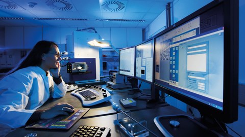 Eine Mitarbeiterin in weißem Laborkittel sitzt in einem kühl beleuchteten physikalischen Labor vor einer Reihe von Monitoren.