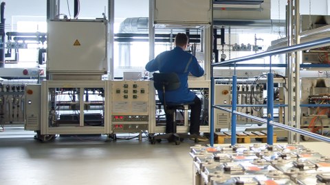 Ein Mann im blauen Arbeitskittel sitzt in einem lichtdurchfluteten Labor voller Rohrleitungen und komplexer technischer Apparaturen.
