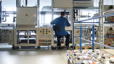 Ein Mann im blauen Arbeitskittel sitzt in einem lichtdurchfluteten Labor voller Rohrleitungen und komplexer technischer Apparaturen.