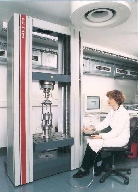 The photograph shows a young woman in a lab coat, sitting in front of the uniaxial pressing device.