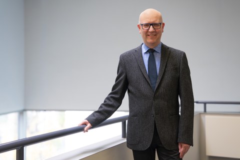 Ein Mann mit Brille und Glatze schaut in die Kamera, während er sich mit der rechten Hand an einem Geländer festhält. Er lacht.