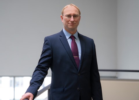 Ein Mann mit blonden Haaren und Brille lehnt an einem Geländer und schaut in die Kamera.