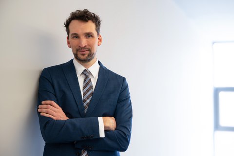 Ein Mann mit lockigen, schwarzen Haaren lehnt an einer Wand und schaut lächelnd in die Kamera.