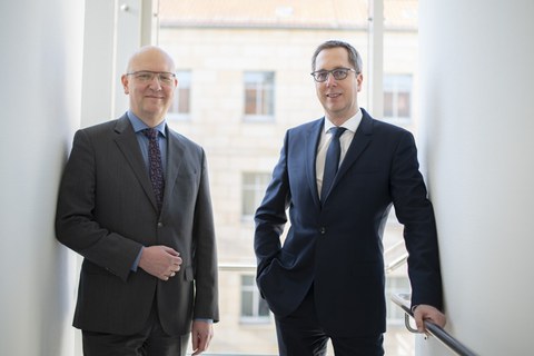 Zwei Männer stehen vor einem Fenster und lächeln in die Kamera.