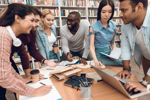 Foto von sechs Studenten aus verschiedenen Ländern, die zusammen in der Bibliothek fröhlich lernen.