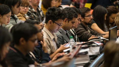 Das Foto zeigt die gut gefüllten Bankreihen in einem Hörsaal. Viele der Studierenden schauen auf ihre Handys oder auf die vor ihn liegenden Taschen.