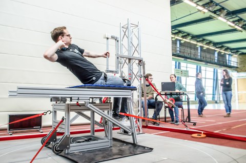 Man sits on a table-like construction, hand resting on the neck as if throwing a ball. Man performs movement. Two people in the background at a laptop.