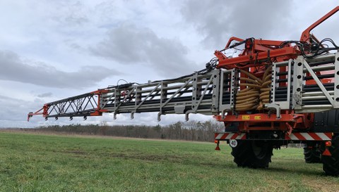 Foto eines Düngemittelstreuers, der an einem Traktor angebracht ist. im Hintergrund: grüne Landschaft und wolkiger Himmel.