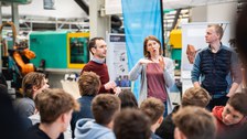 A group of young people in the foreground listen attentively to a man and a woman. The woman is speaking sign language and gesticulating. A second man stands at the left edge of the picture. In the background: machines.