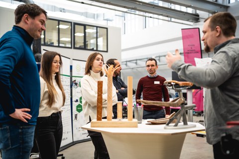 A group of young people and men stand in front of a table on which demonstrators have been set up. A man gesticulates and explains the demonstrators on the table.