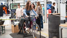 A girl sits on a bicycle and pedals - students stand in the background, which is dominated by machines and screens in a large hall.