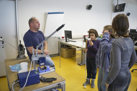 Right: A technician sits on a table next to the HoliPress and explains something to three schoolgirls in the right part of the picture. One pupil is blind and has a cane in her hand.