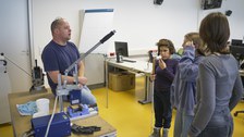 Right: A technician sits on a table next to the HoliPress and explains something to three schoolgirls in the right part of the picture. One pupil is blind and has a cane in her hand.