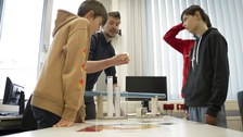 An engineer is standing around a table with three boys. On the table are various plastics in tubular containers. The boys look and listen attentively.