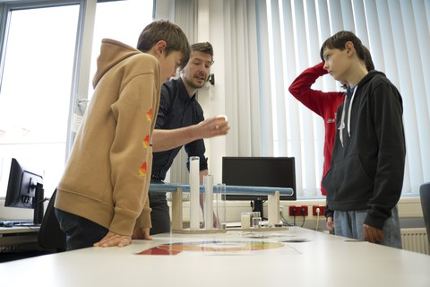 An engineer is standing around a table with three boys. On the table are various plastics in tubular containers. The boys look and listen attentively.