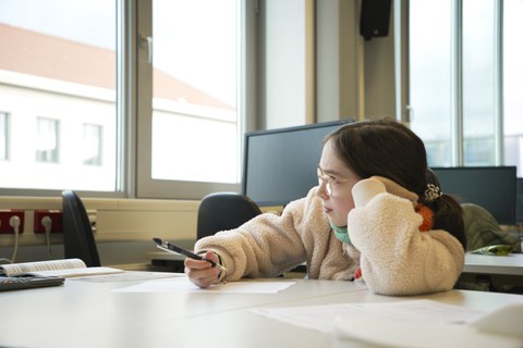 A girl is sitting at a table, one elbow resting on the table. She holds a pen in her other hand and a sheet of paper in front of her. The girl is looking intently out of the window, as if she is thinking hard.