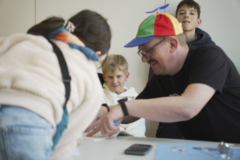 Zwei Jungen schauen schelmisch in die Kamera. Im Vordergrund (leicht unscharf) erklärt ein Techniker einem Mädchen etwas auf einem Blatt Papier und lächelt.