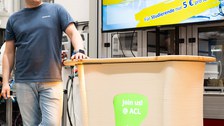 Eine Person steht angelehnt an ein Podest. Ein im Podest integrierter Bildschirm zeigt die Aufschrift "Join us @ ACL" an.