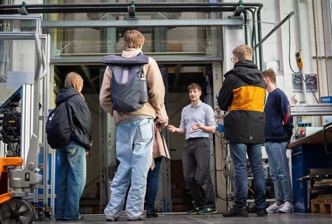 Ein ILK-Mitarbeiter erklärt einer Gruppe von Kindern ein technisches Exponat. Im Hintergrund ist der Fallturm im Leichtbau- und Innovationszentrum (LIZ) zu sehen.