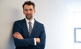 Porttrait eines Mannes mit schwarzen Locken und blauem Anzug. Er lehnt gegen eine Wand und lächelt freundlich in die Kamera.