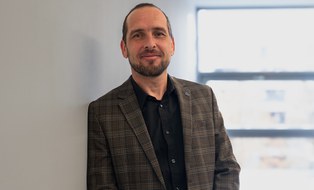 A man wearing a plaid suit and with black hair leans against a wall and looks calmly into the camera.