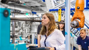 A student stands at a braiding machine, holding a display device while looking toward the machine as if inspecting it. In the background, other students and a technician observe her.