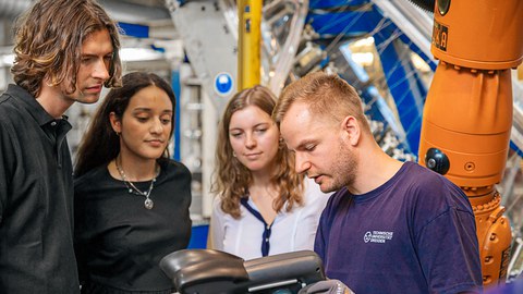 Ein Mann erklärt einer Gruppe von Student:innen in einem technischen Umfeld ein Bauteil, das er mit Handschuhen in der Hand hält. Die Student:innen stehen im Halbkreis um ihn, daneben befindet sich ein Roboterarm.