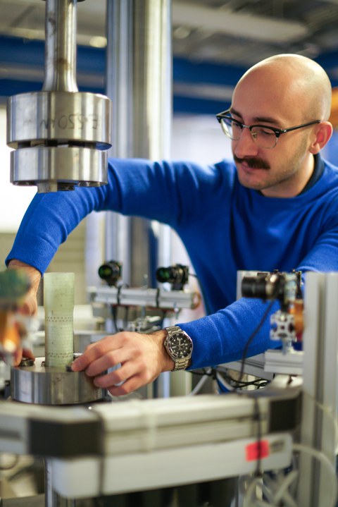 Ein Mann mit Schnurrbart, Brille und Glatze beugt sich über einen Prüfstand, um ein Bauteil einzuspannen.