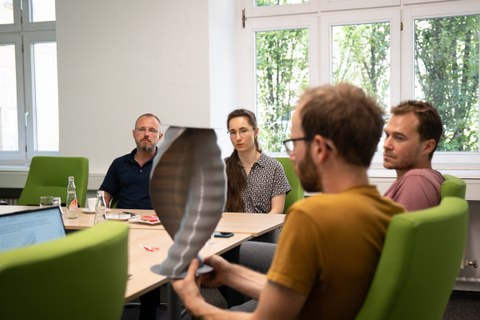 Personen sitzend an einem Tisch. Im Vordergrund hält eine Person ein Bauteil (unscharf), im Hintergrund ist die junge Wissenschaftlerin zu sehen (scharf).