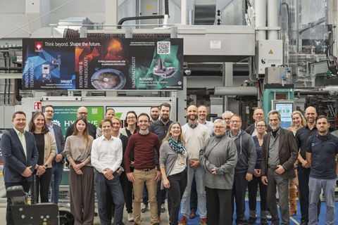 Das Gruppenfoto zeigt eine Gruppe von Personen, die vor einer groß dimensionierten Maschine stehen und in die Kamera blicken. Dabei ist eine positive, lachende Gestik zu beobachten.