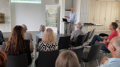 Prof. Dr. Modler im Vortrag, im Vordergrund sitzen die Gäst:innen, die dem Vortragenden lauschen. Im Hintergrund: eine auf die Wand projizierte PowerPoint-Präsentation.