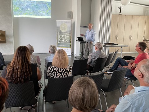 Prof. Dr. Modler im Vortrag, im Vordergrund sitzen die Gäst:innen, die dem Vortragenden lauschen. Im Hintergrund: eine auf die Wand projizierte PowerPoint-Präsentation.