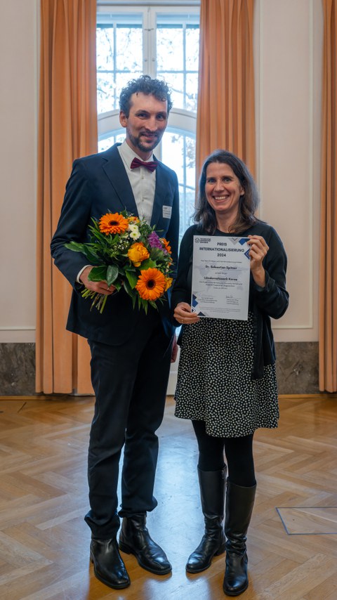 Zwei Personen, eine Frau und ein Mann, lächeln in die Kamera und halten eine Urkunde hoch.