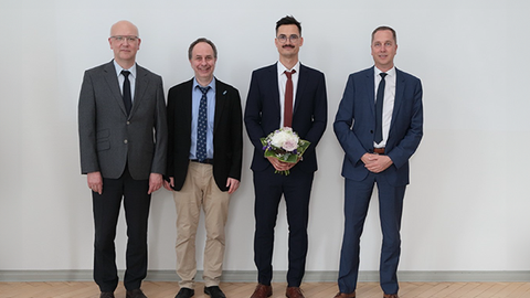 Vier Personen stehen in einer Reihe und schauen in die Kamera. Der Promovend hält einen Blumenstraß in der Hand.