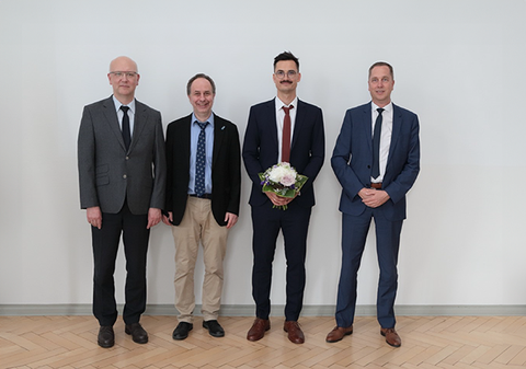 Four people stand in a row and look into the camera. The doctoral candidate is holding a bouquet of flowers.