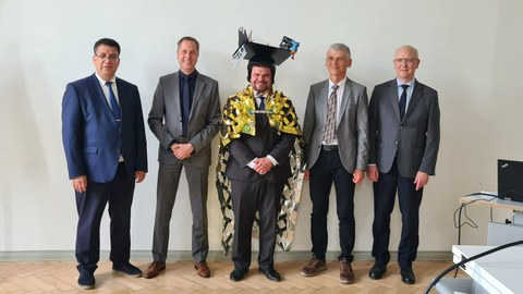 The men are standing next to each other and smiling at the camera. The man in the centre is wearing a doctor's hat.
