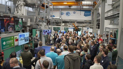 In der Halle des LEIV stehen viele Personen in einem Halbkreis um eine Maschine. Ein Mann steht mit einem Mikrofon vor einem Bildschirm und erklärt.