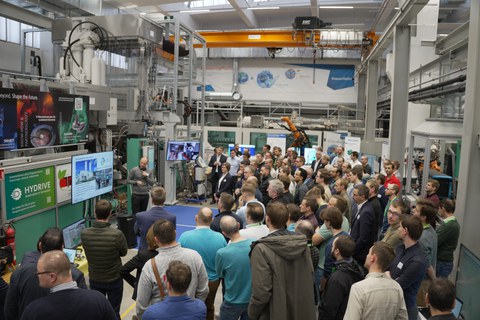 In der Halle des LEIV stehen viele Personen in einem Halbkreis um eine Maschine. Ein Mann steht mit einem Mikrofon vor einem Bildschirm und erklärt.